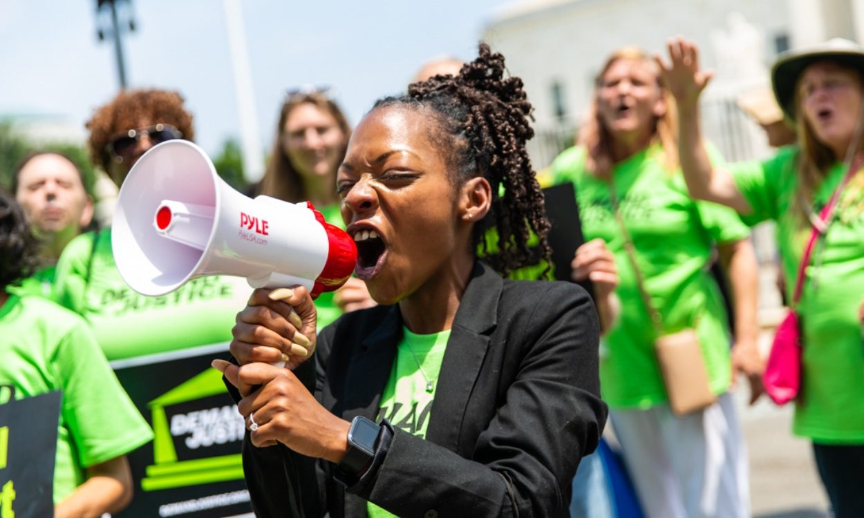 image of woman screaming into bullhorn in the foreground with demand justice volunteers wearing green shirts standing behind her